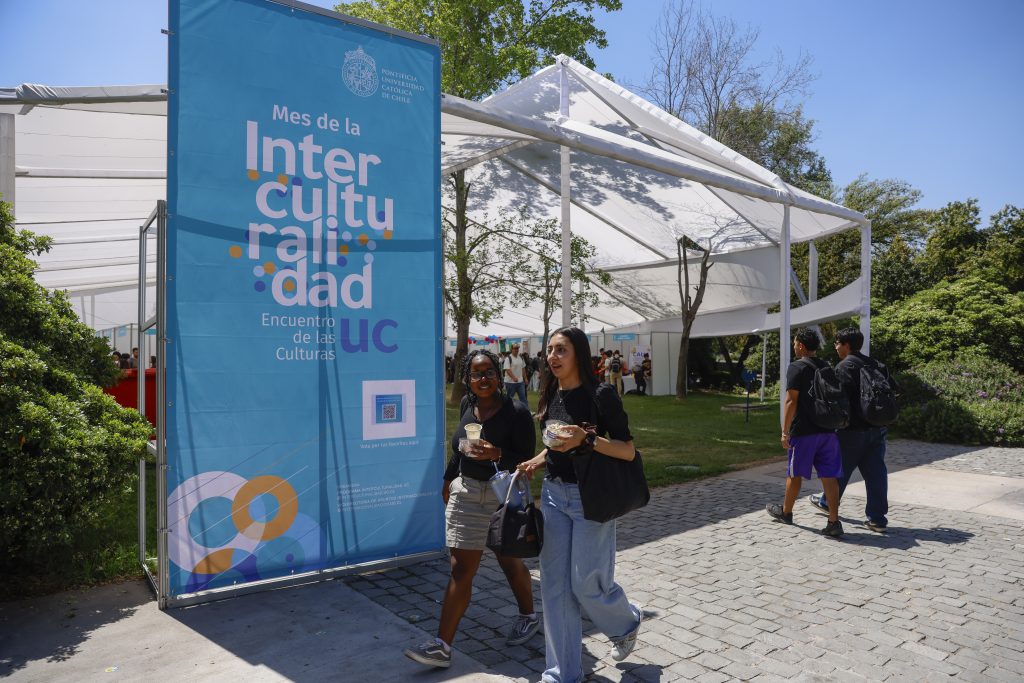 Dos estudiantes mujeres caminan por la arboleda del Campus San Joaquín, frente al letrero de Interculturalidad UC.